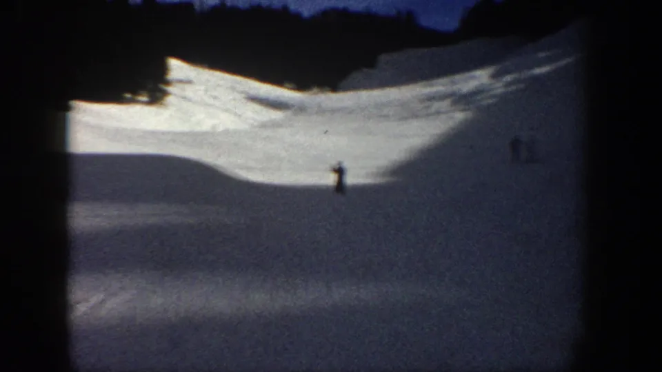 1961 Aspen home-movie frame with a distant skier on a broad snowy slope, dark trees, and blue evening light.