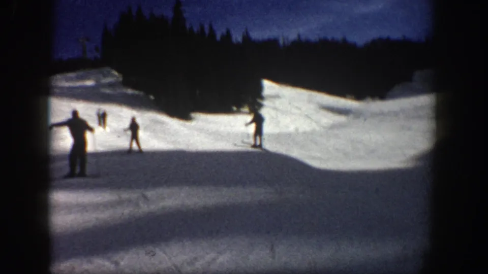 1961 Aspen home-movie frame showing distant skiers on a snowy slope bordered by dark evergreens and a deep blue sky.