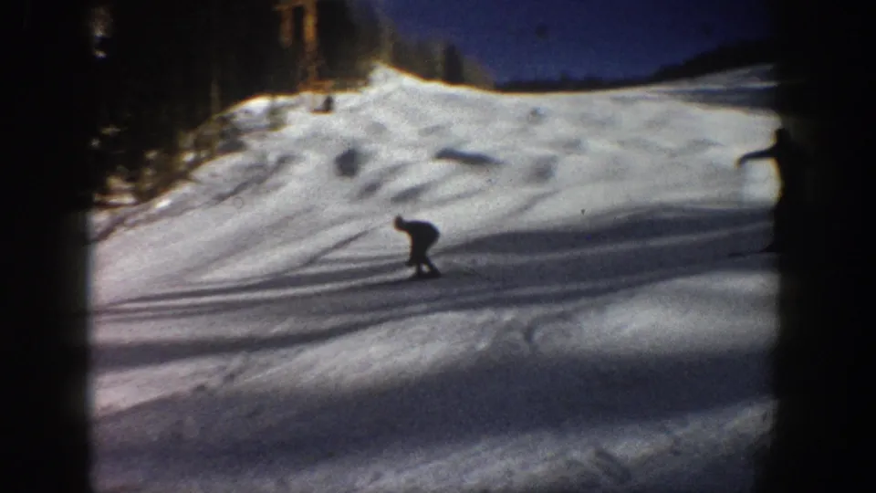 Distant skier descends an Aspen snow slope in 1961 home-movie footage, framed by trees and dark 8mm gate edges.