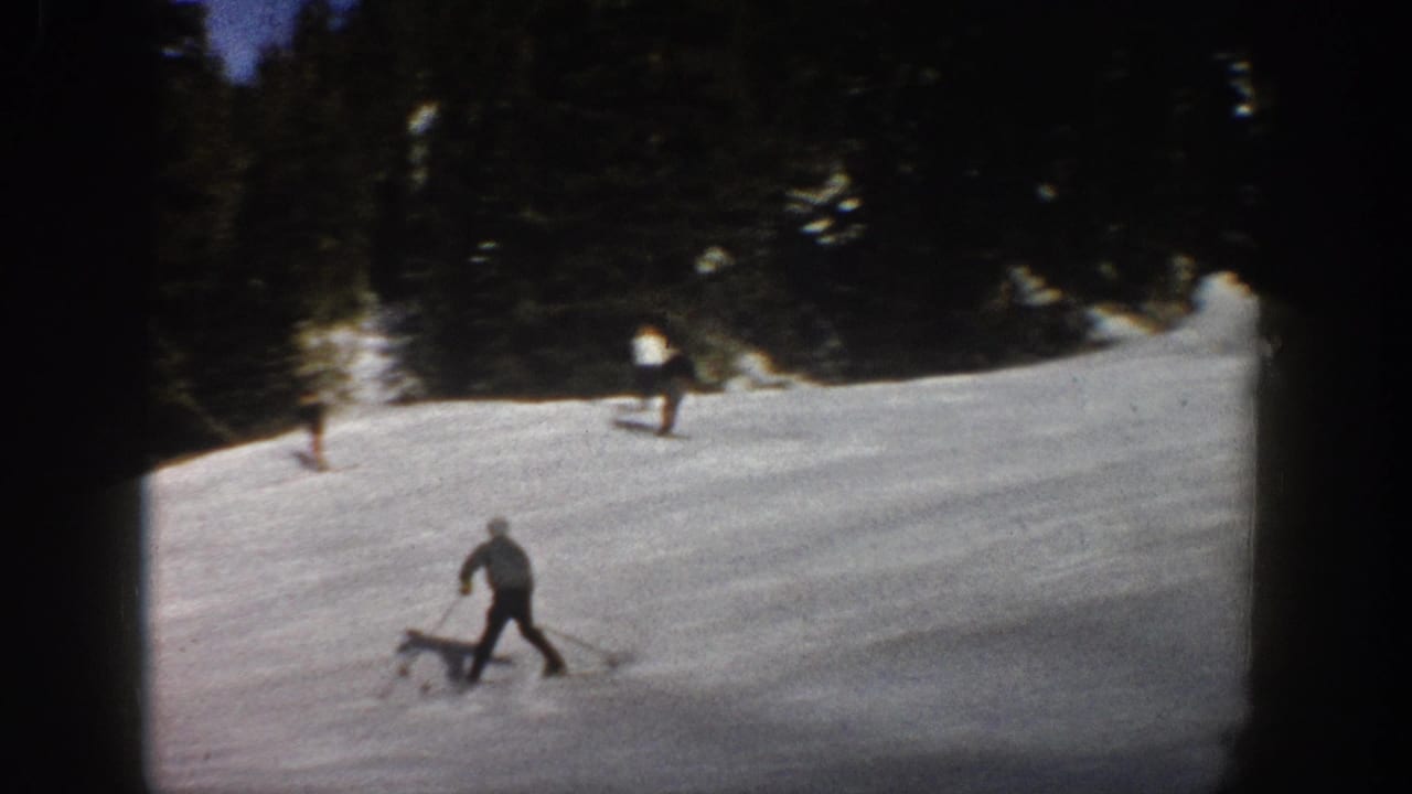 Aspen 1961 home-movie frame showing skiers spread across a snowy run below a dark evergreen tree line.