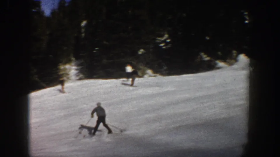 Aspen 1961 home-movie frame showing skiers spread across a snowy run below a dark evergreen tree line.