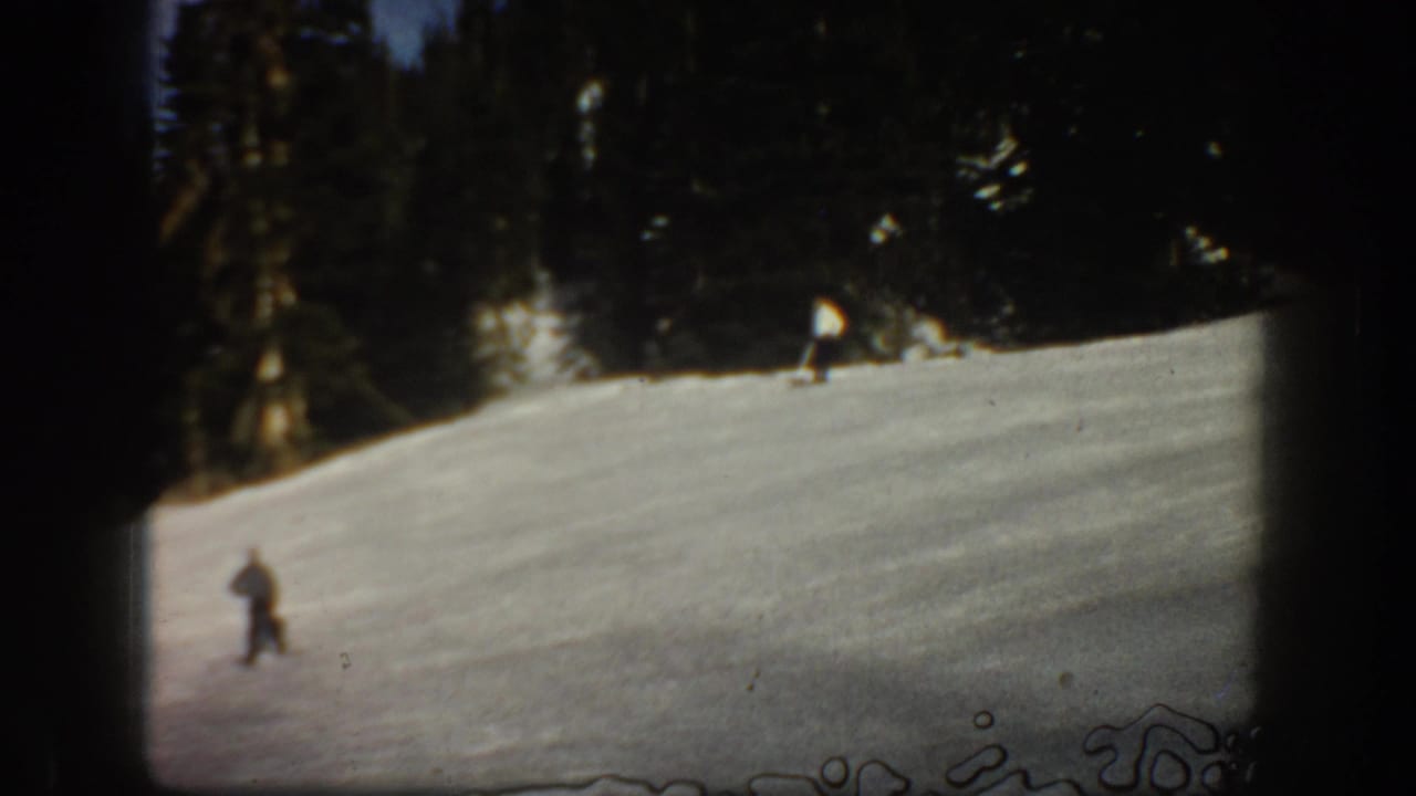1961 Aspen frame with a distant skier moving across a bright snowy run under a dense evergreen backdrop.