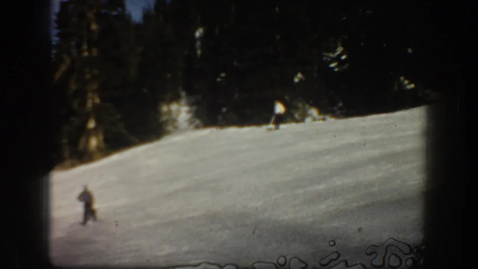 1961 Aspen frame with a distant skier moving across a bright snowy run under a dense evergreen backdrop.