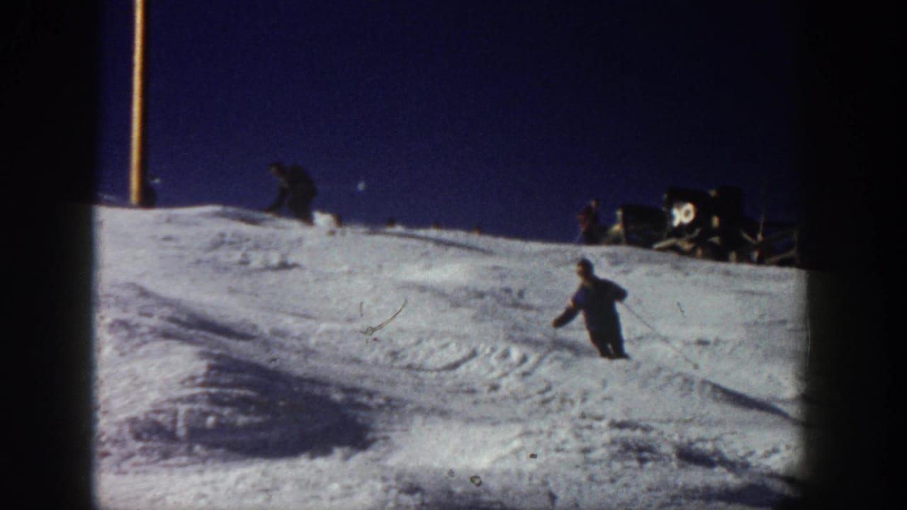 1961 Aspen night-skiing frame showing a lit snowy slope, a skier in motion, a vertical pole, and people near dark equipment.