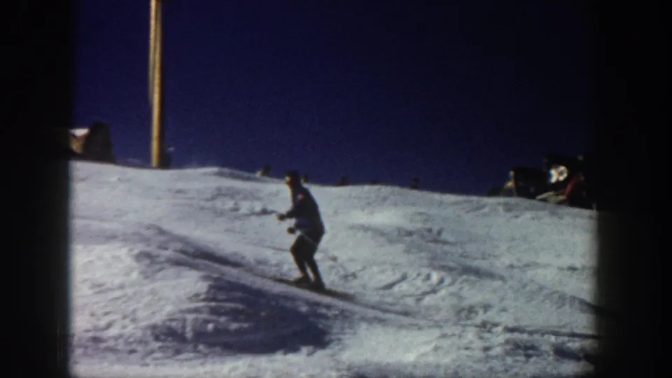 1961 Aspen home-movie frame showing a skier turning on a lit night slope with a pole and dark sky behind.