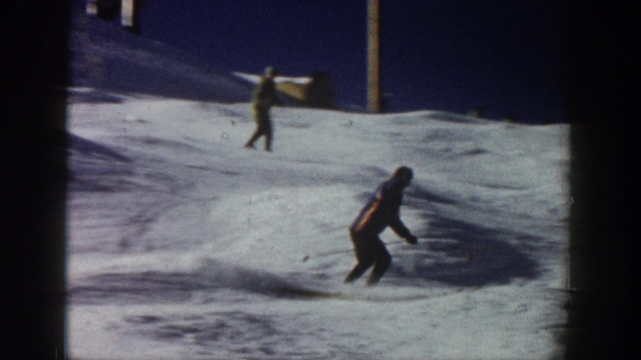 Two skiers appear on a snowy slope, one closer in dark clothing and another in the background.