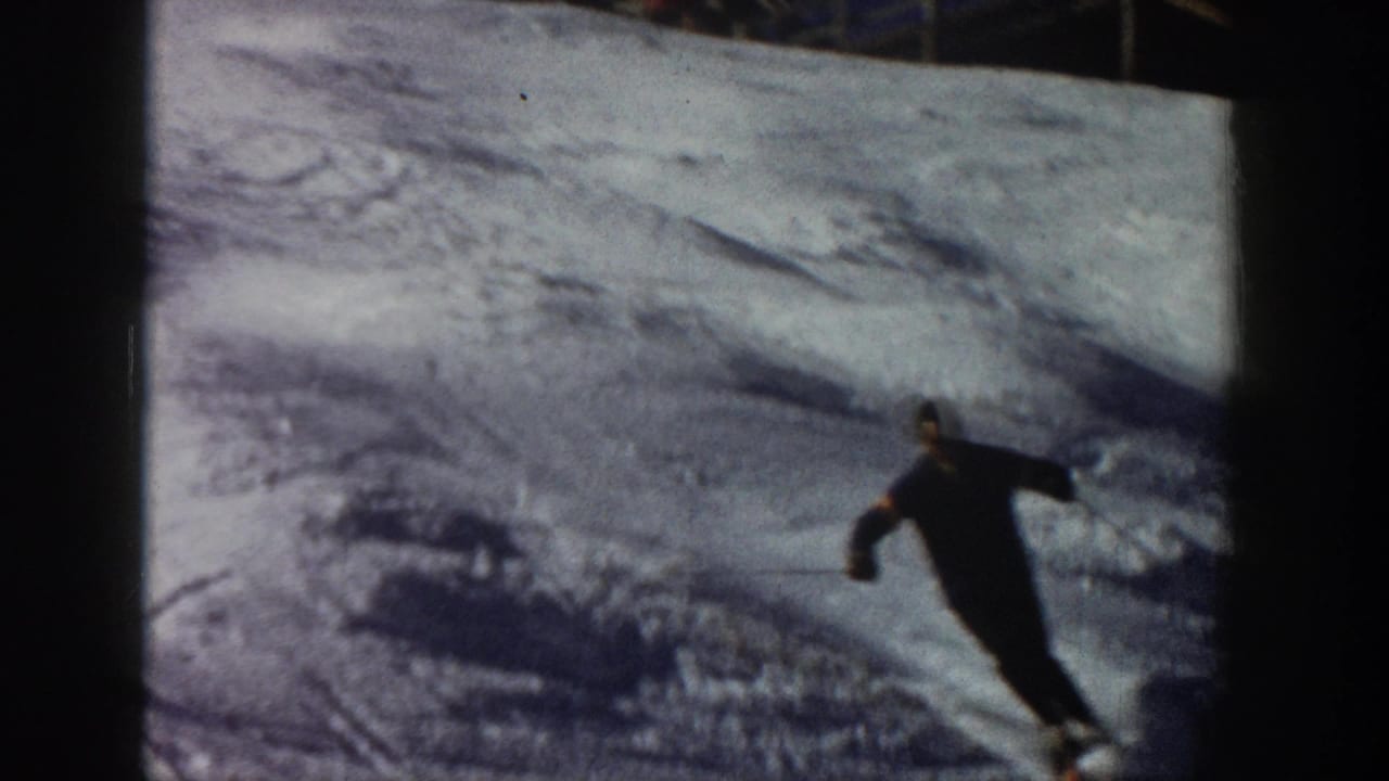 1961 Aspen home-movie frame showing a skier in dark clothing carving through uneven snow with poles visible.