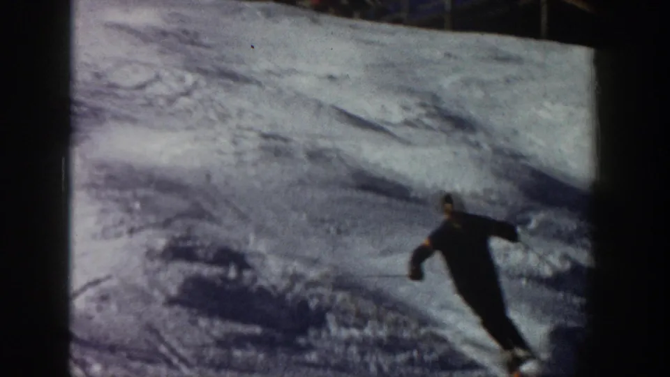 1961 Aspen home-movie frame showing a skier in dark clothing carving through uneven snow with poles visible.