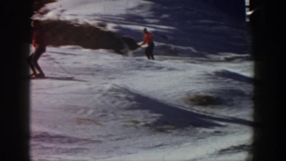 Aspen, Colorado 1961 home-movie frame with one skier entering at left and another in red crossing a snowy slope near exposed ground.