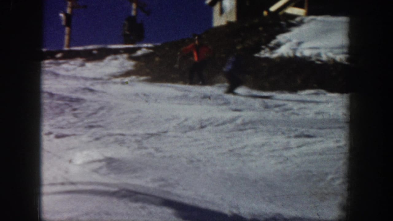 Aspen, Colorado 1961 frame showing a distant red-clad skier on a snow slope, with poles, exposed ground, and part of a small structure nearby.