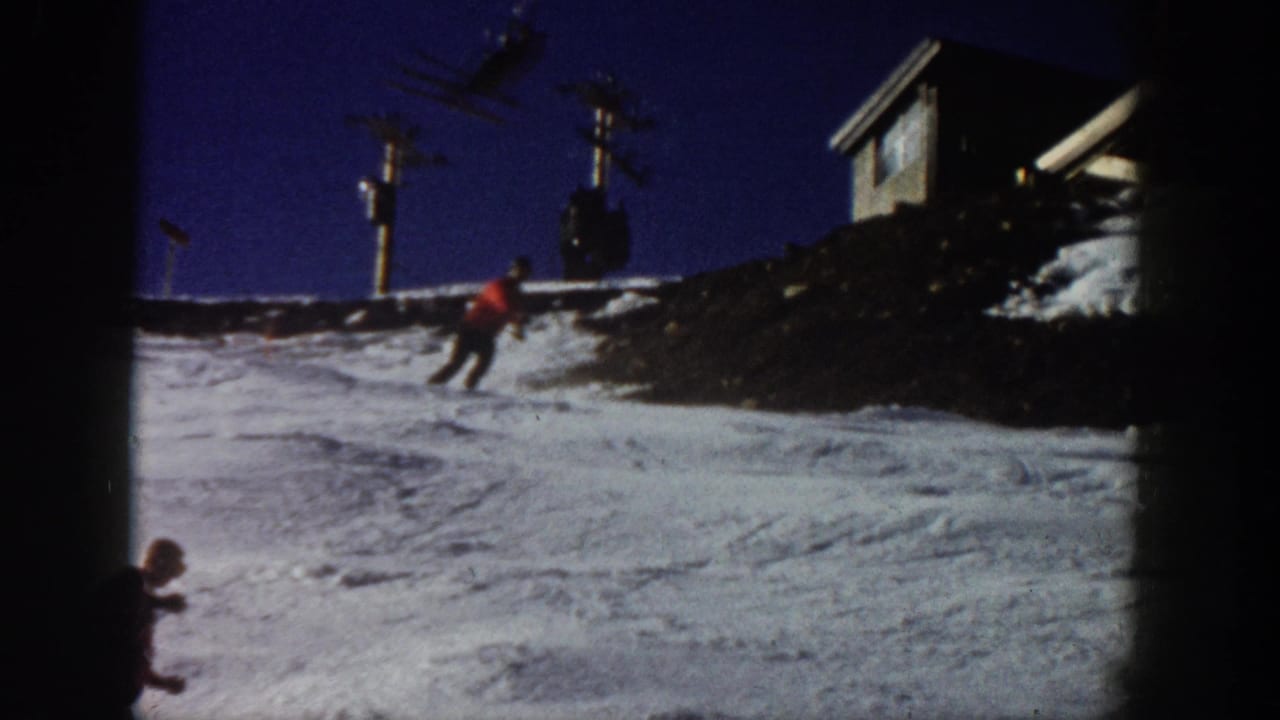 1961 Aspen home-movie frame showing a red-jacketed skier descending a snowy slope near lift infrastructure and a lodge-like structure.