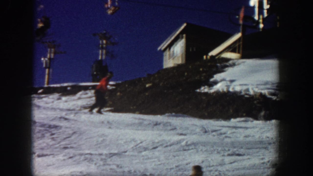 Red-jacketed skier continues downhill near a snowy embankment and lift structures.
