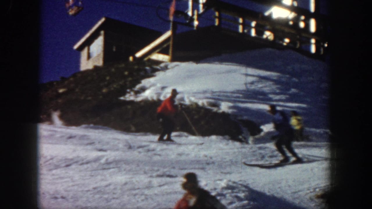 1961 Aspen home-movie frame showing multiple skiers on a slope, including a red-jacketed skier near a lodge-like building.