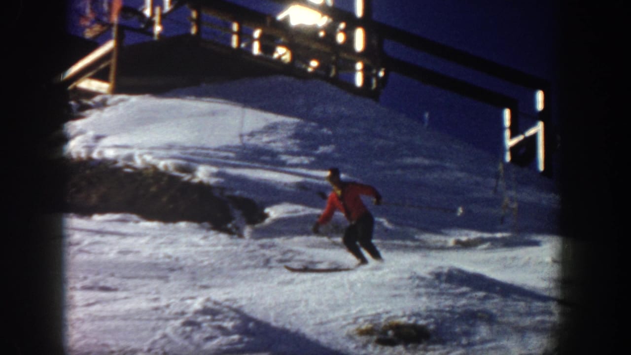 In Aspen, Colorado, 1961, a skier in a red jacket begins descending a snowy slope with a lift structure overhead.