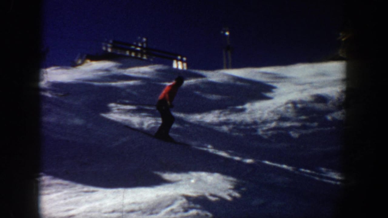 An adult skier appears on a lit snowy slope in Aspen, Colorado, with dark sky and bright artificial lights in the distance.