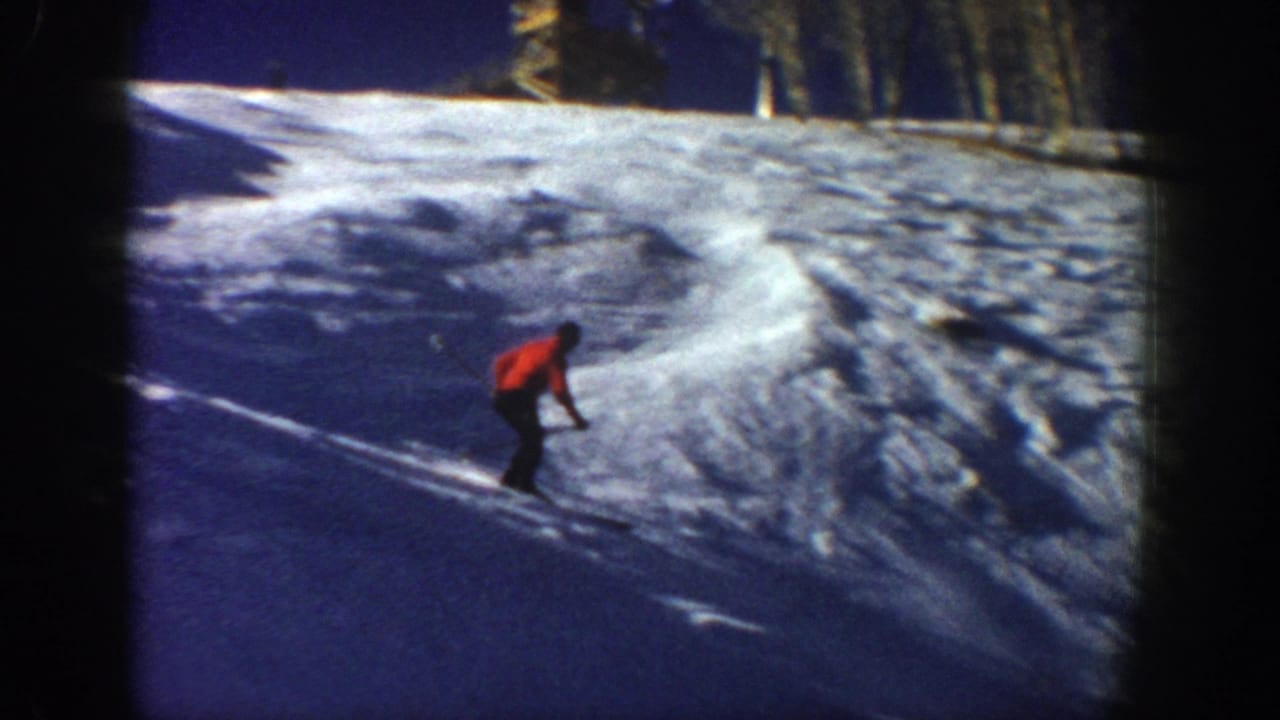 The home-movie frame shows the adult skier leaning into a descent on a snow-covered Aspen slope, with trees near the upper edge.