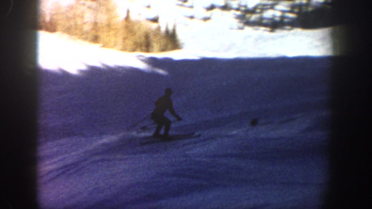A distant skier continues downhill on a shaded Aspen slope, with pine trees and bright snow visible along the upper edge.