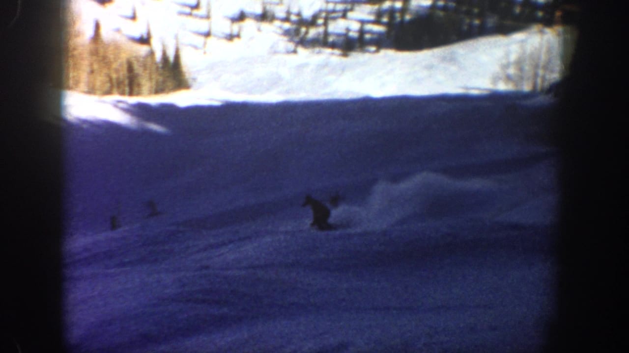 1961 Aspen home-movie frame showing a small skier silhouette on a steep blue-shadowed slope below bright snow and evergreen trees.