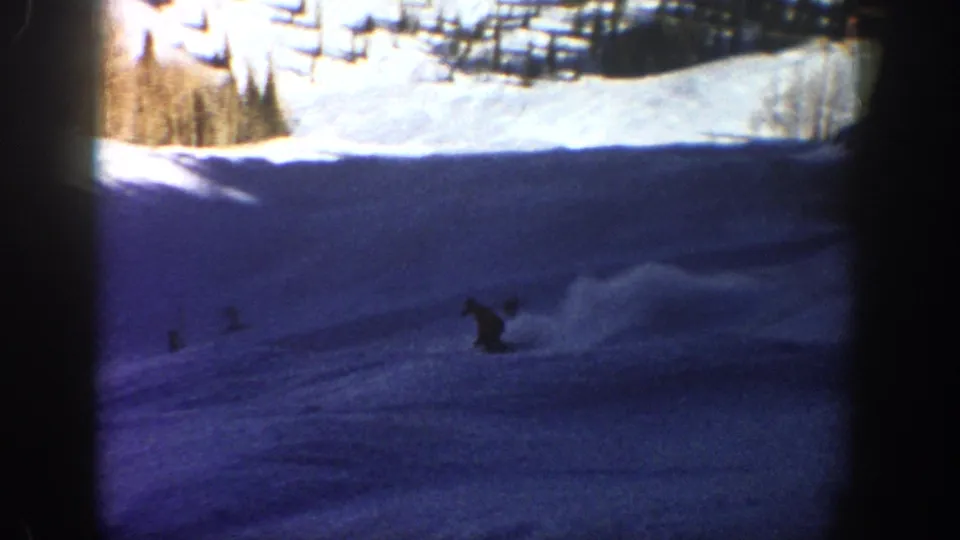 1961 Aspen home-movie frame showing a small skier silhouette on a steep blue-shadowed slope below bright snow and evergreen trees.