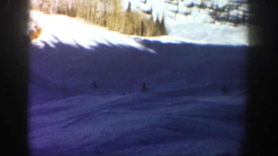 1961 Aspen 8mm frame of a broad shadowed ski slope with small distant figures and sunlit evergreens along the upper edge.