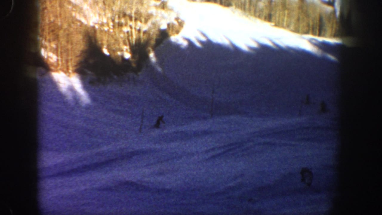 1961 Aspen 8mm frame showing distant skier silhouettes on a shadowed mountain run, with sunlit snowbanks and trees above.