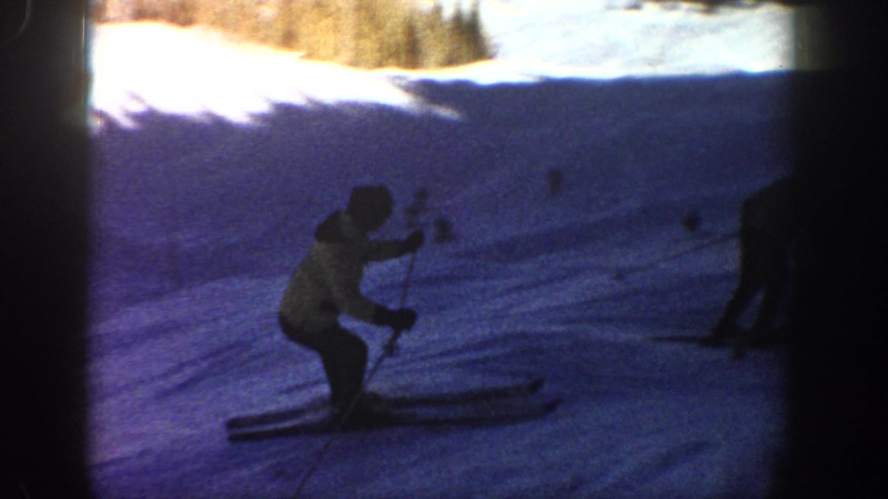 1961 Aspen home-movie frame with a skier in a yellow jacket crossing a shadowed section of snowy terrain.