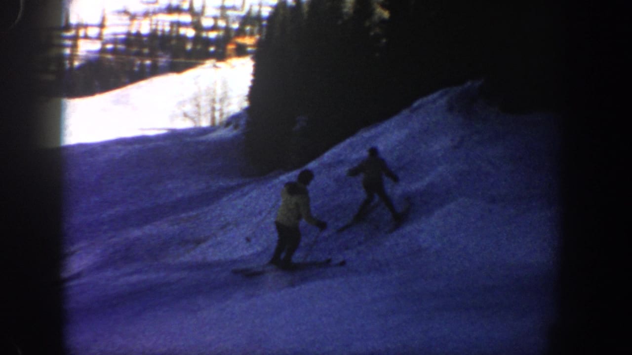 1961 Aspen home-movie frame showing two skiers on a shaded snowy slope with forest and fence shapes in the background.