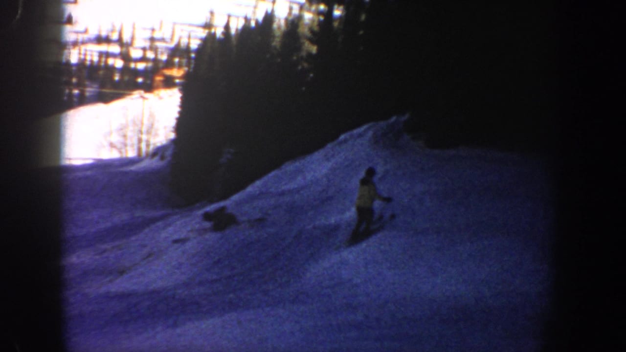 A skier stands on a snowy slope while another person lies or falls nearby.
