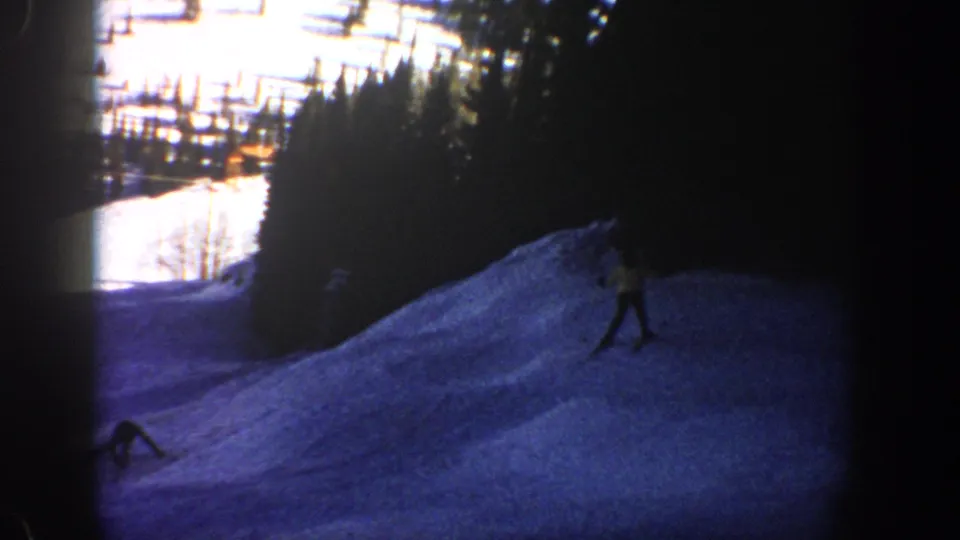 1961 Aspen home-movie frame with a skier moving across a blue-tinted snowy slope bordered by dense trees.