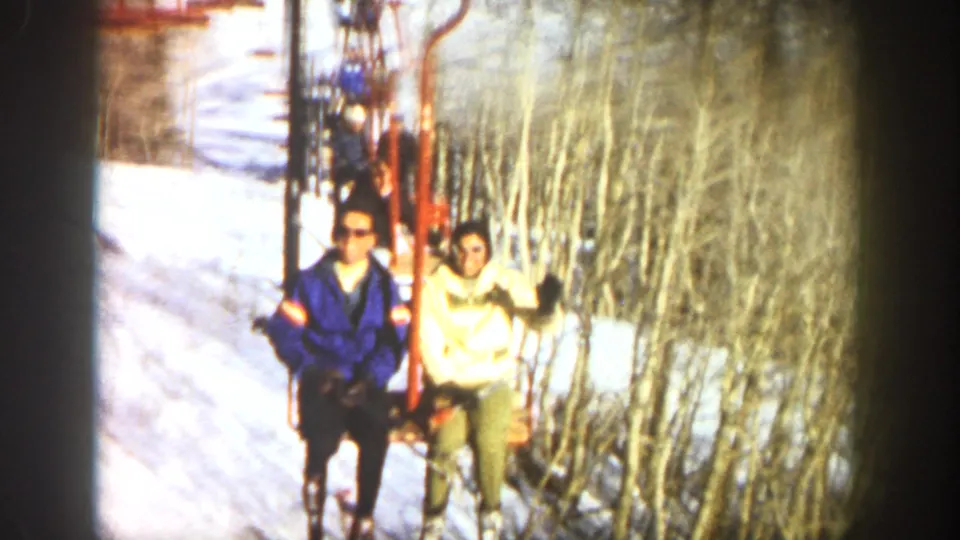 1961 Aspen frame showing two visible riders seated on a ski lift passing through snow and leafless trees.