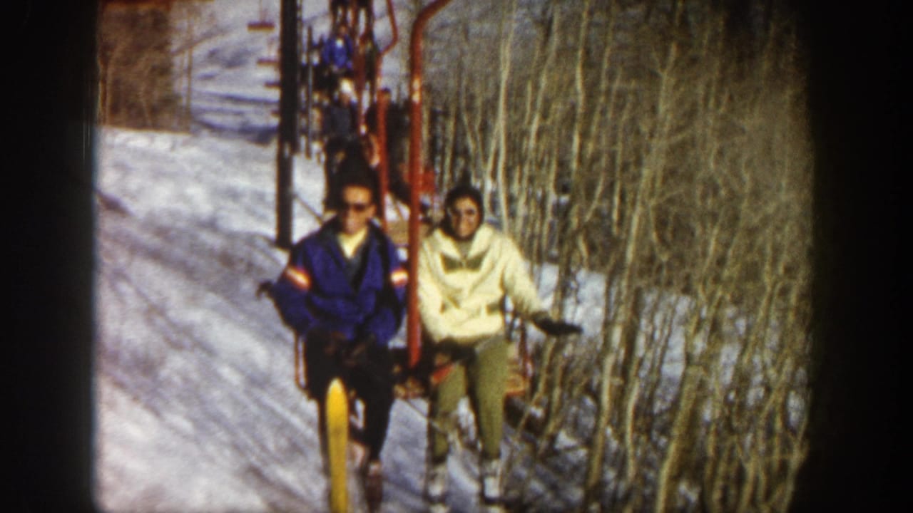 Two adult skiers on a red chairlift in Aspen, Colorado, with snow and bare aspen trees around them.