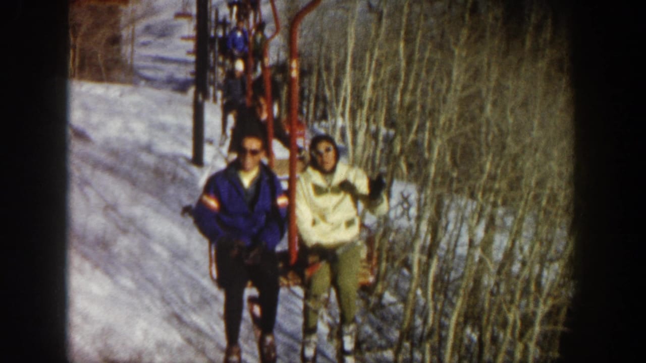 Two skiers face the camera from a chairlift, with one waving beside snowy trees.