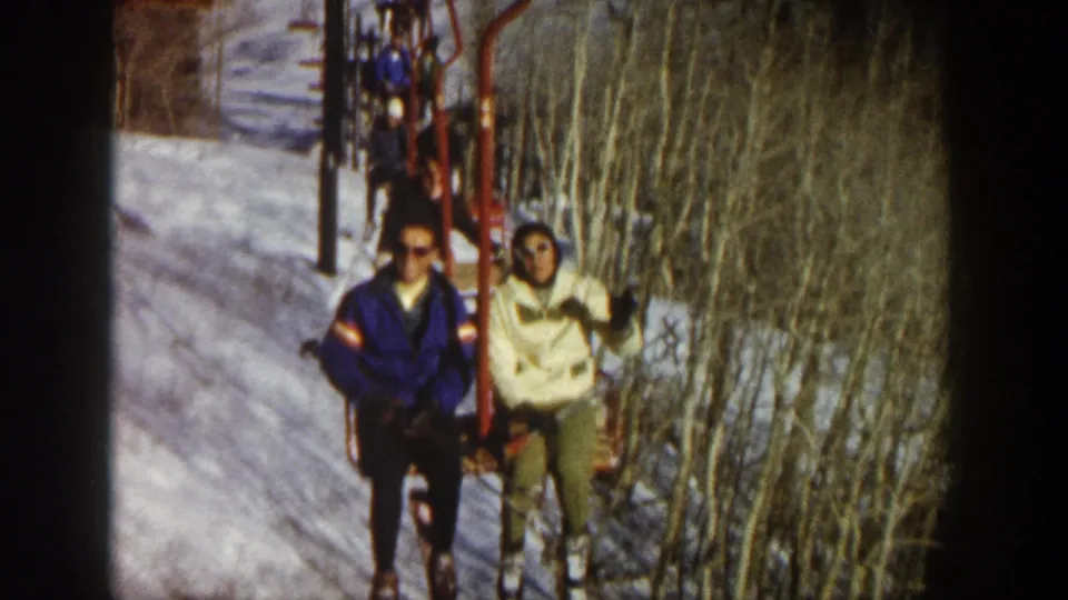 In the 1961 Aspen home-movie footage, two skiers ride a red chairlift as the woman waves toward the camera.