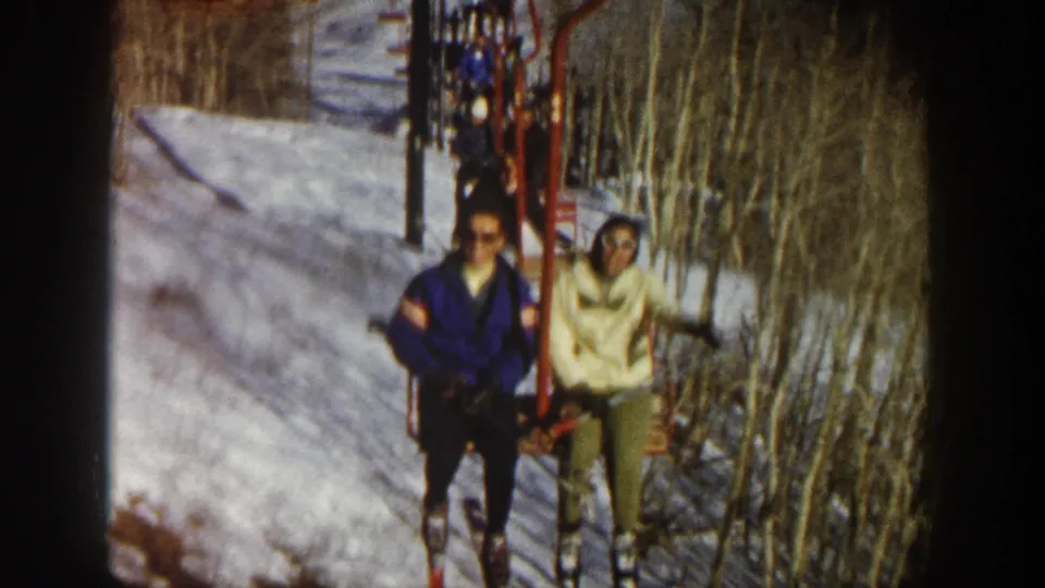 Two adult skiers continue upward on a red chairlift in Aspen, with snowy ground and bare aspen trunks visible behind them.
