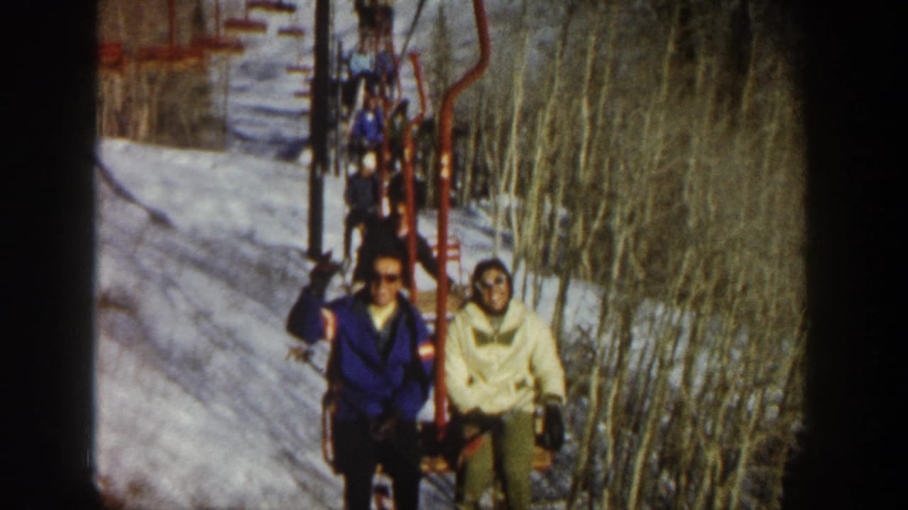 Two people in 1960s ski clothing ride a red chairlift in Aspen, Colorado, with snowy ground and bare trees around the lift line.