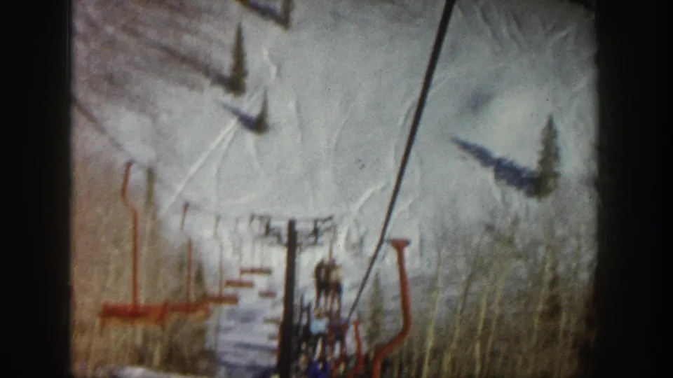 A red chairlift and cable line pass over a snowy Aspen slope, with lift shadows, trees, and a distant seated rider visible.