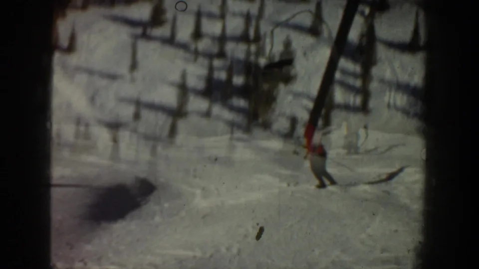 A skier descends a snowy Aspen slope below the chairlift, with strong shadows, lift structures, and trees visible in the background.
