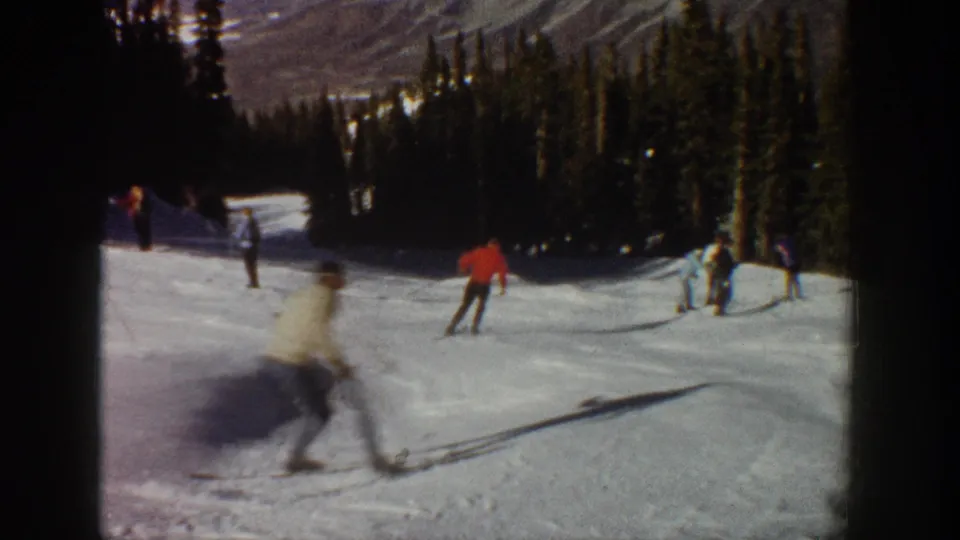 Several skiers move down a broad Aspen ski run bordered by evergreens, with mountainside terrain visible in the distance.