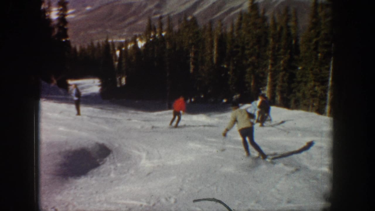 1961 Aspen home-movie frame showing several skiers moving downhill on a snow-covered slope with evergreens and mountains beyond.