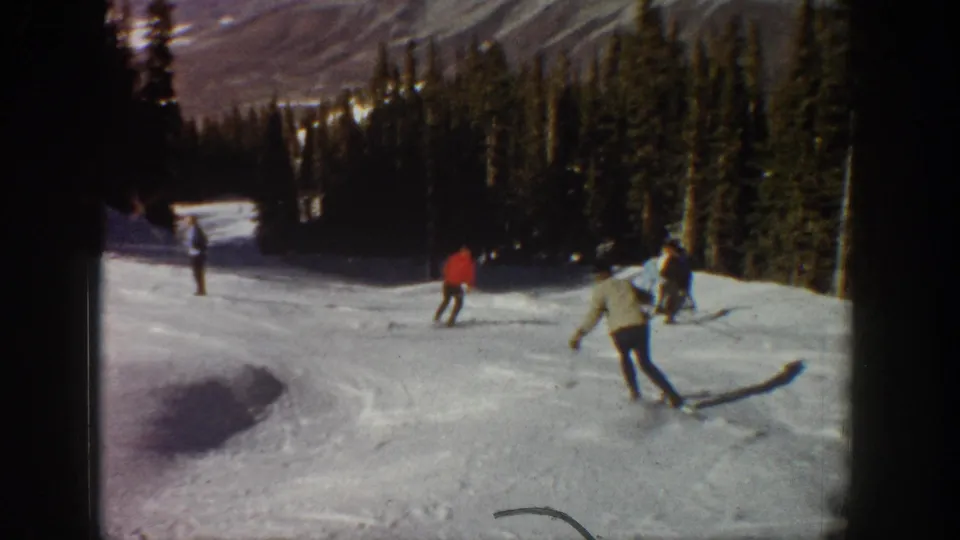 1961 Aspen home-movie frame showing several skiers moving downhill on a snow-covered slope with evergreens and mountains beyond.