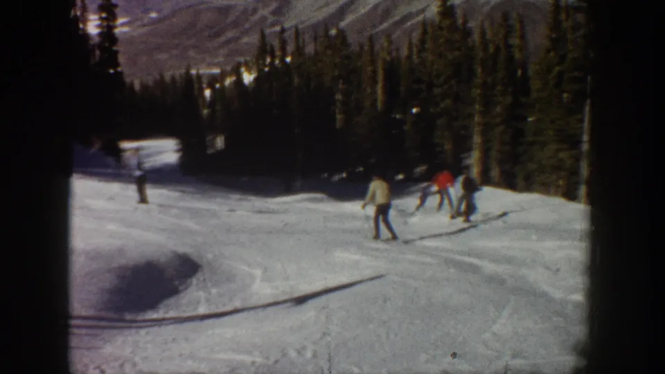 1961 Aspen ski footage frame with skiers in winter jackets crossing a bright snowy run beside dark evergreen trees.