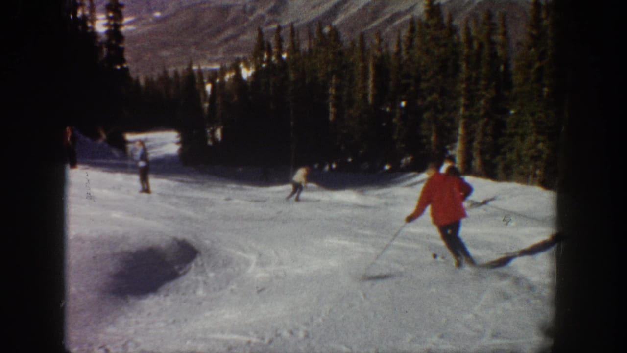 1961 Aspen home-movie frame showing a red-jacketed skier turning downhill while other skiers continue across the snow.