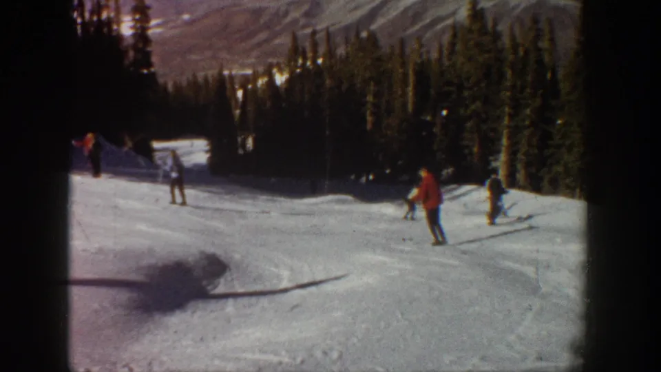 1961 Aspen frame showing skiers spaced across a snowy mountain run, with evergreen forest and distant slopes in the background.