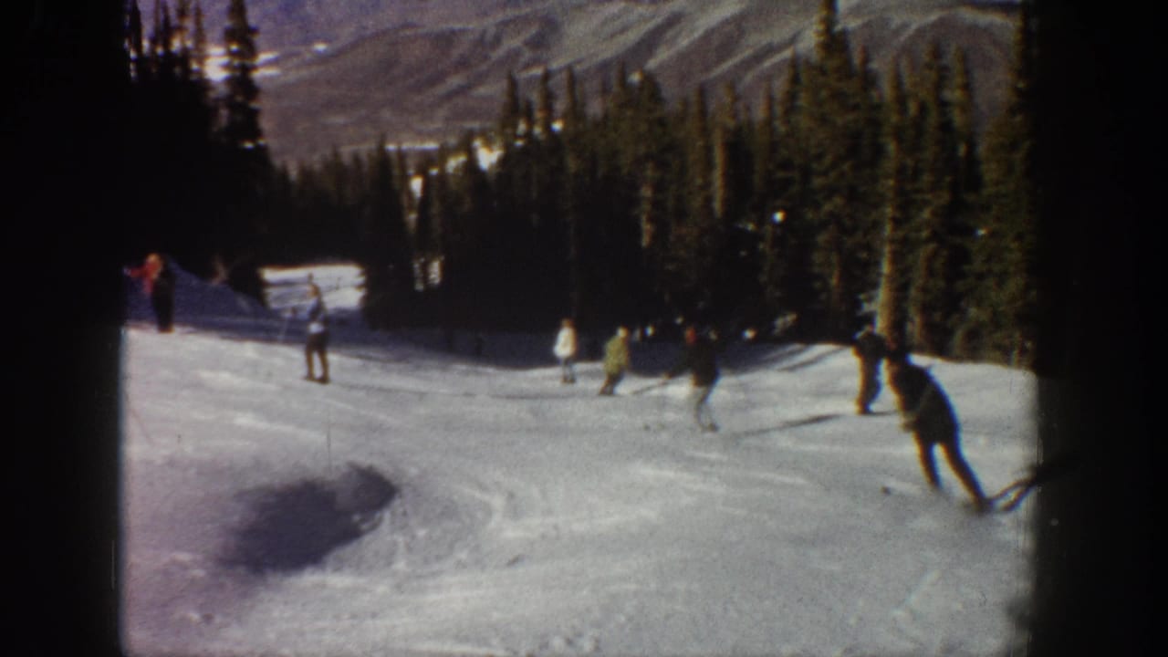 Skiers descend a snowy slope between evergreens with mountains in the background.