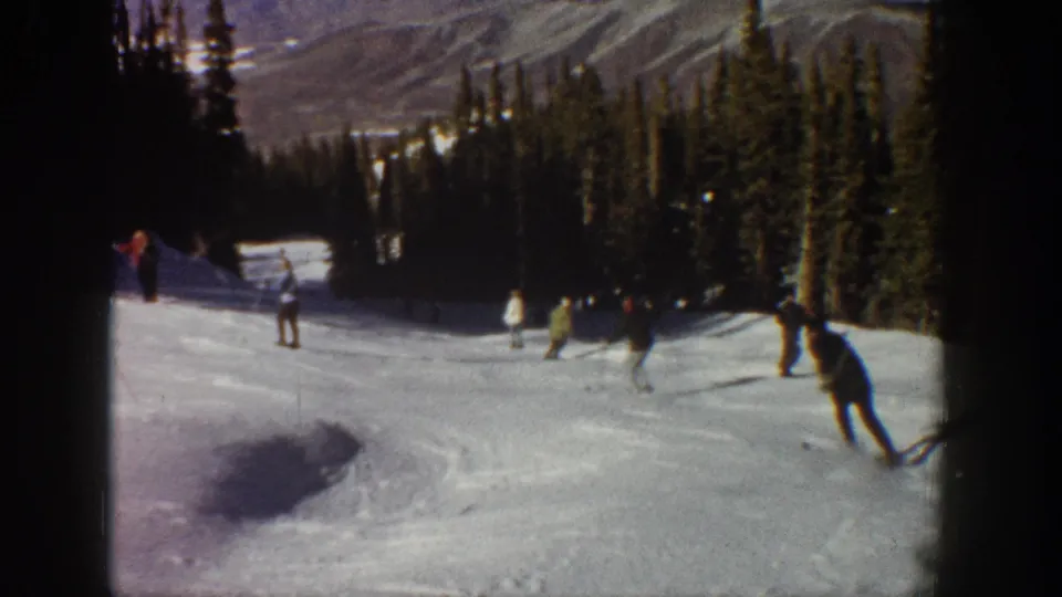 1961 Aspen-area home-movie frame showing several skiers moving down a snowy run bordered by evergreens and distant mountains.