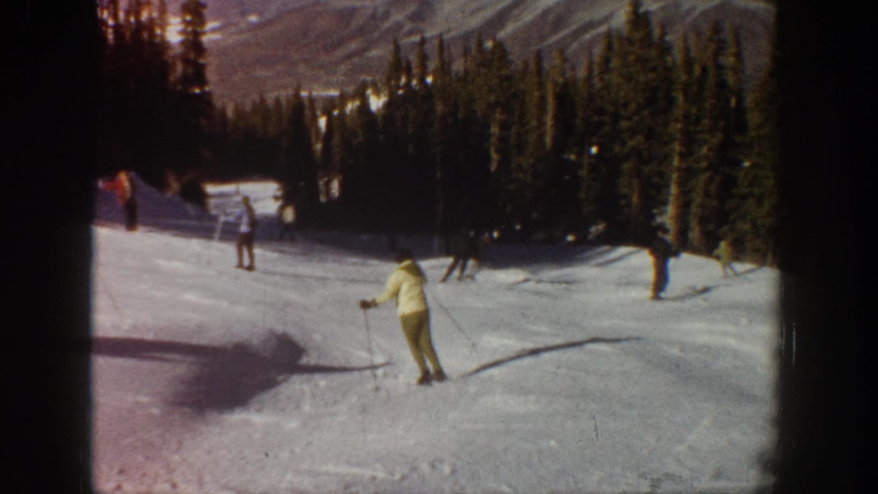 1961 Aspen-area home-movie frame showing a light-colored skier carving across the lower run while distant skiers pass near the trees.