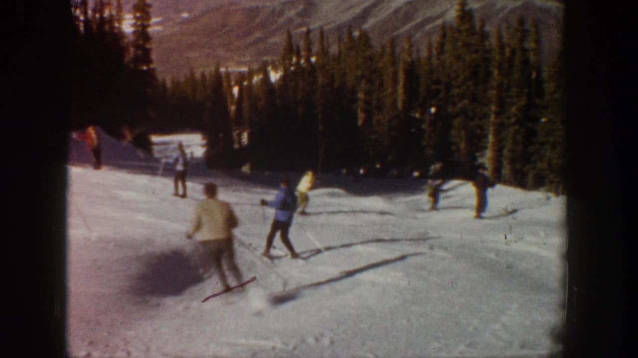 1961 Aspen home-movie frame showing several skiers on a snowy slope with evergreen trees and distant mountains.
