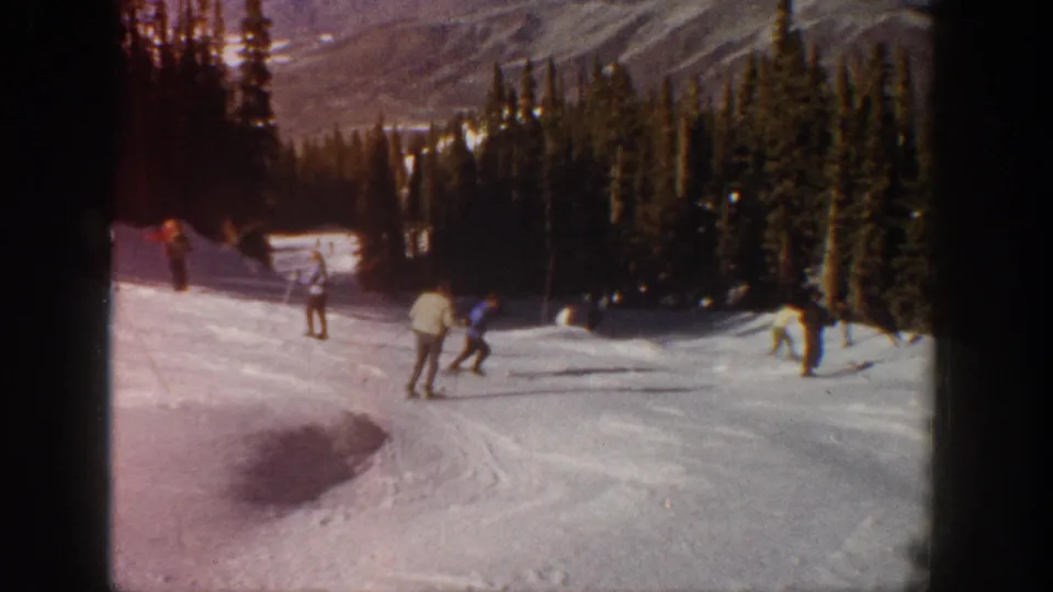 1961 Aspen ski vacation frame with skiers spread across a snowy run below dense evergreens and mountain ridges.