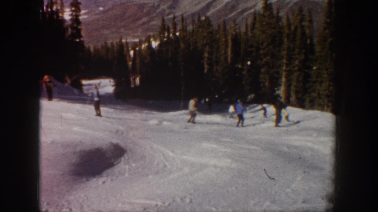 Home-movie frame from Aspen in 1961 showing distant skiers pausing and gliding on a snow-covered slope near forest.