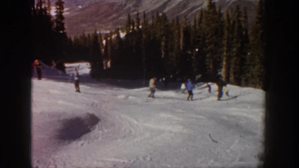 Home-movie frame from Aspen in 1961 showing distant skiers pausing and gliding on a snow-covered slope near forest.